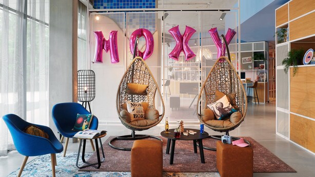 Een modern interieur met twee blauwe stoelen, twee hangende eierstoelen, een klein rond tafeltje en een groot raam met uitzicht.