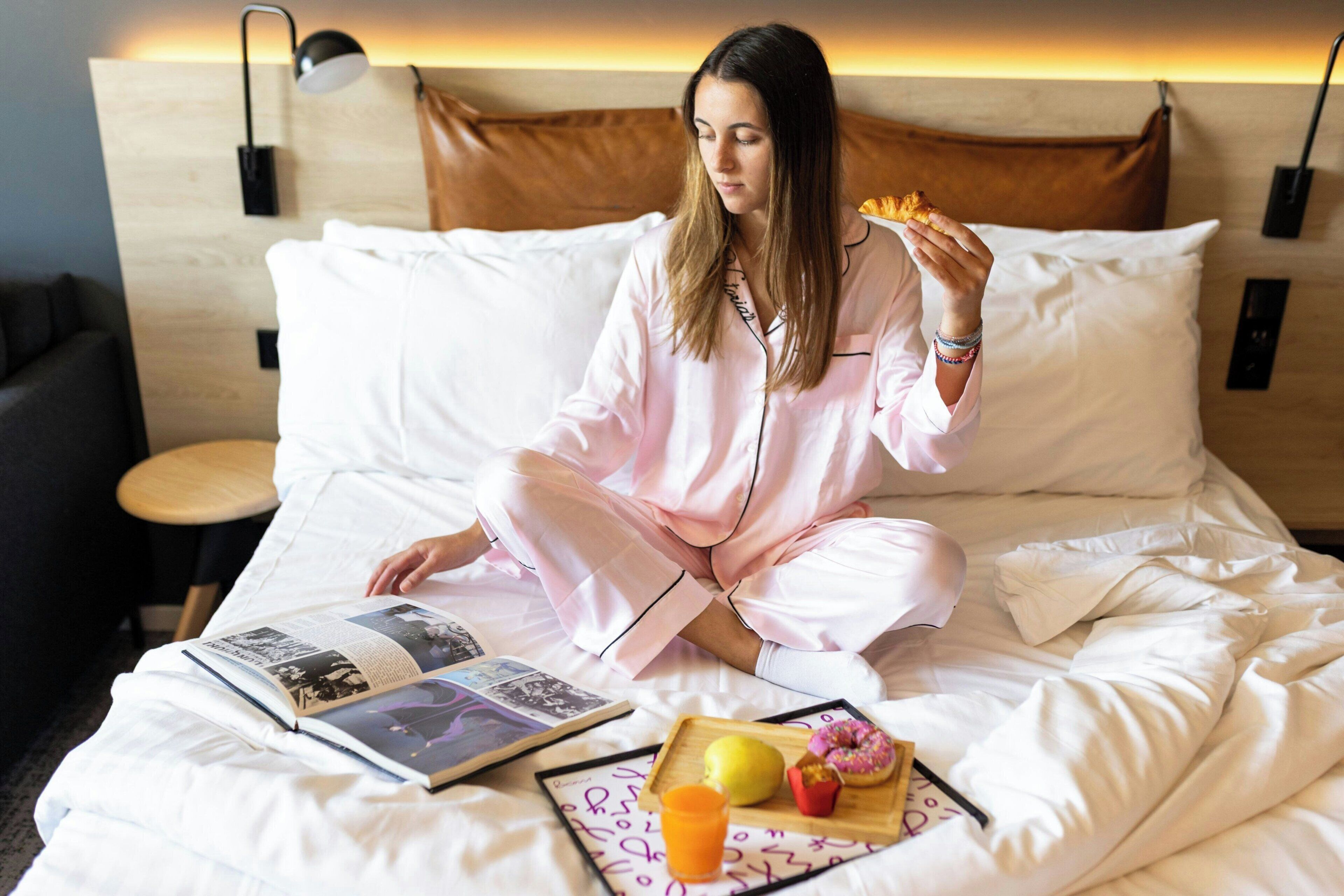 A person sitting on a bed with a book, a tray with food and a drink, and a pillow.