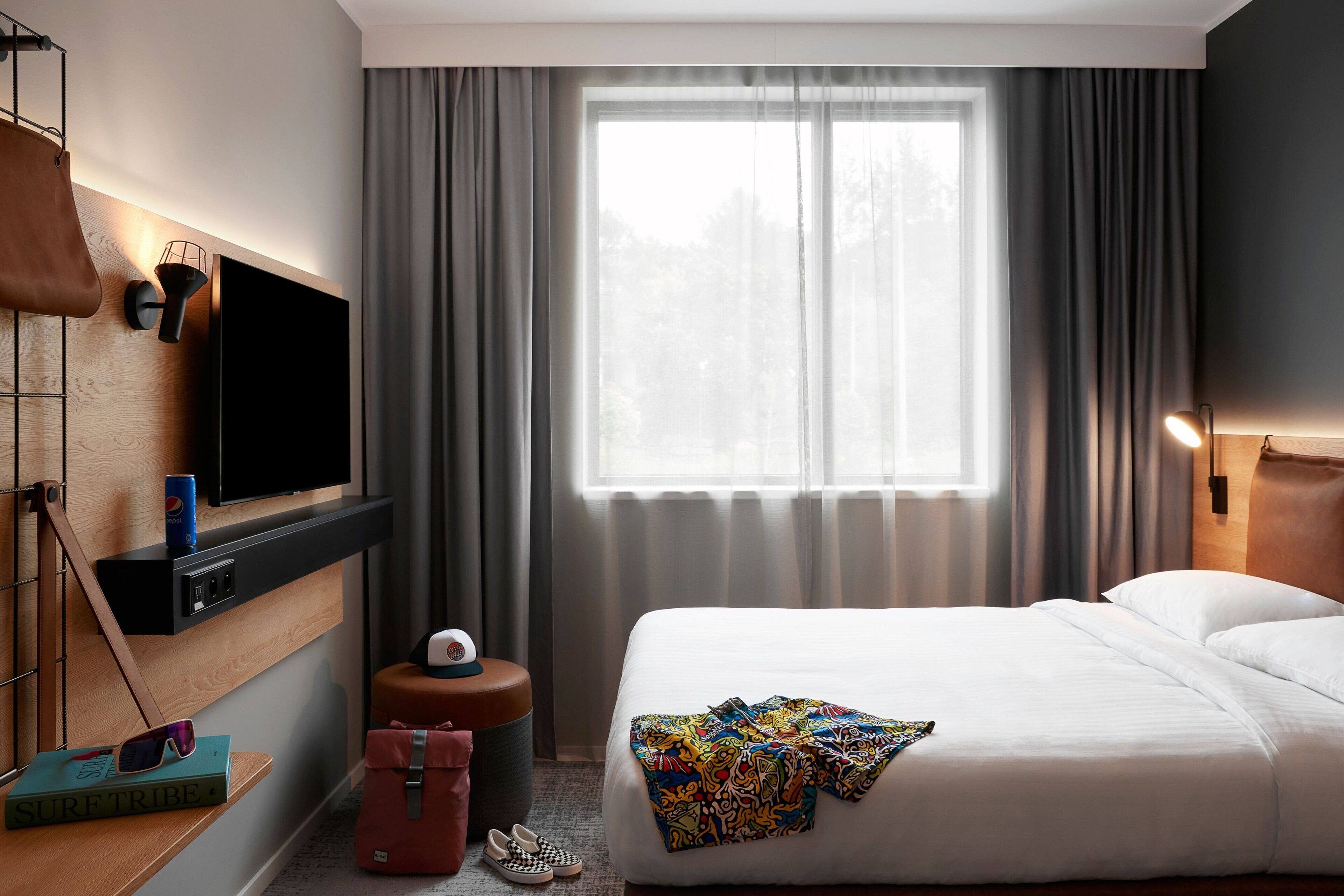 A hotel room with a bed, a TV mounted on the wall, a window with curtains, a small table with a book and sunglasses, and a suitcase on the floor.