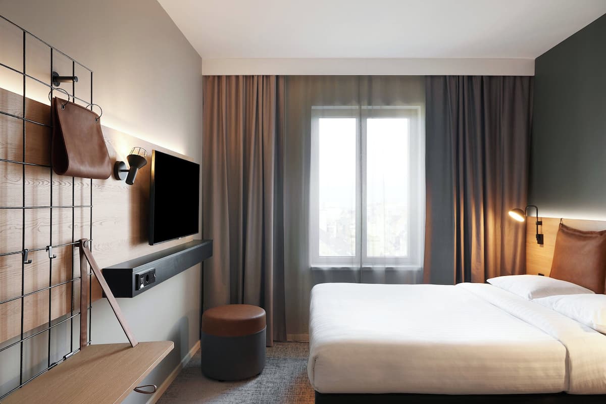 A modern hotel room with a large bed, a flat-screen TV mounted on a wooden panel, a small round ottoman, and a window with curtains.