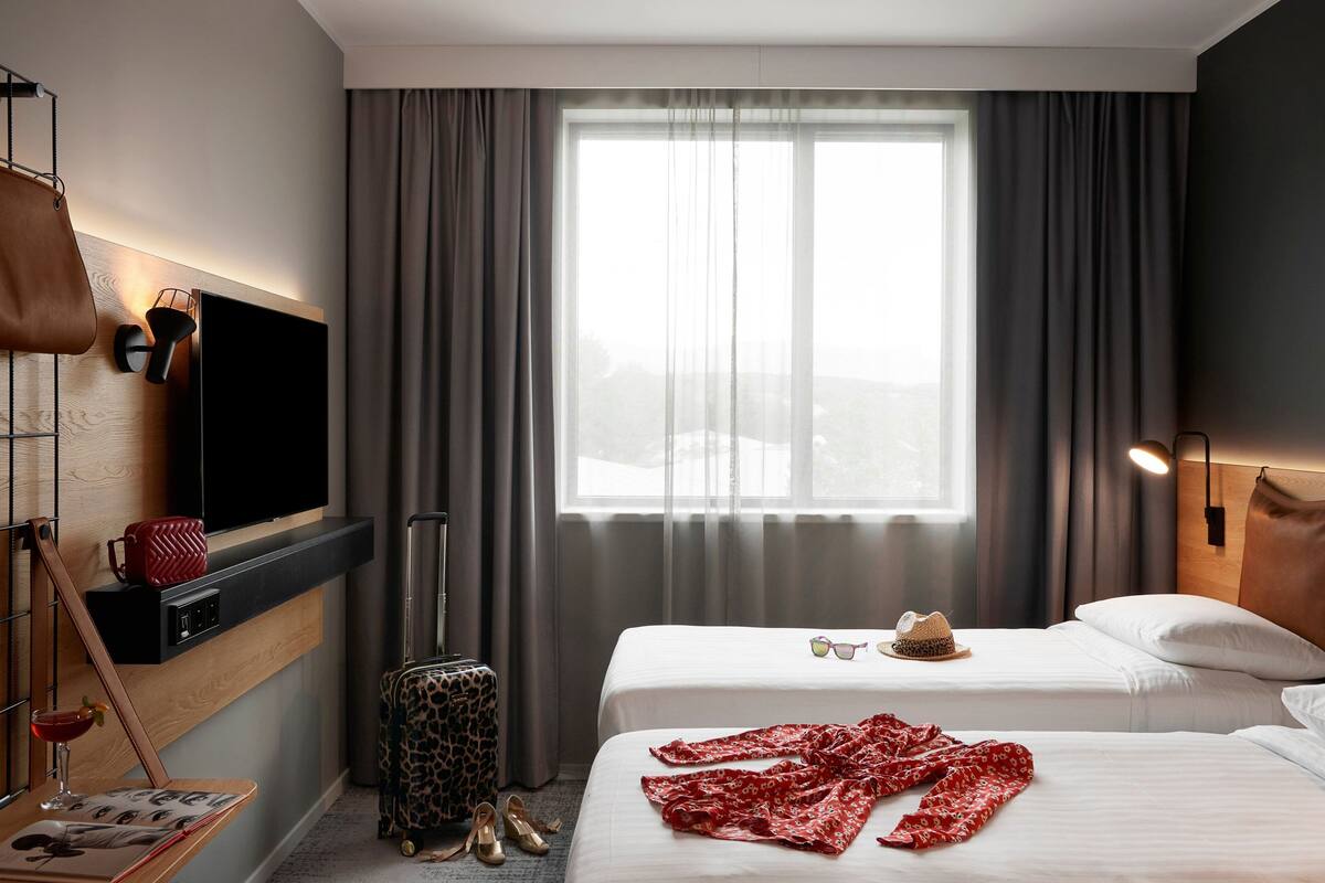 A hotel room with two beds, a TV mounted on the wall, a window with curtains, a suitcase, and a handbag.