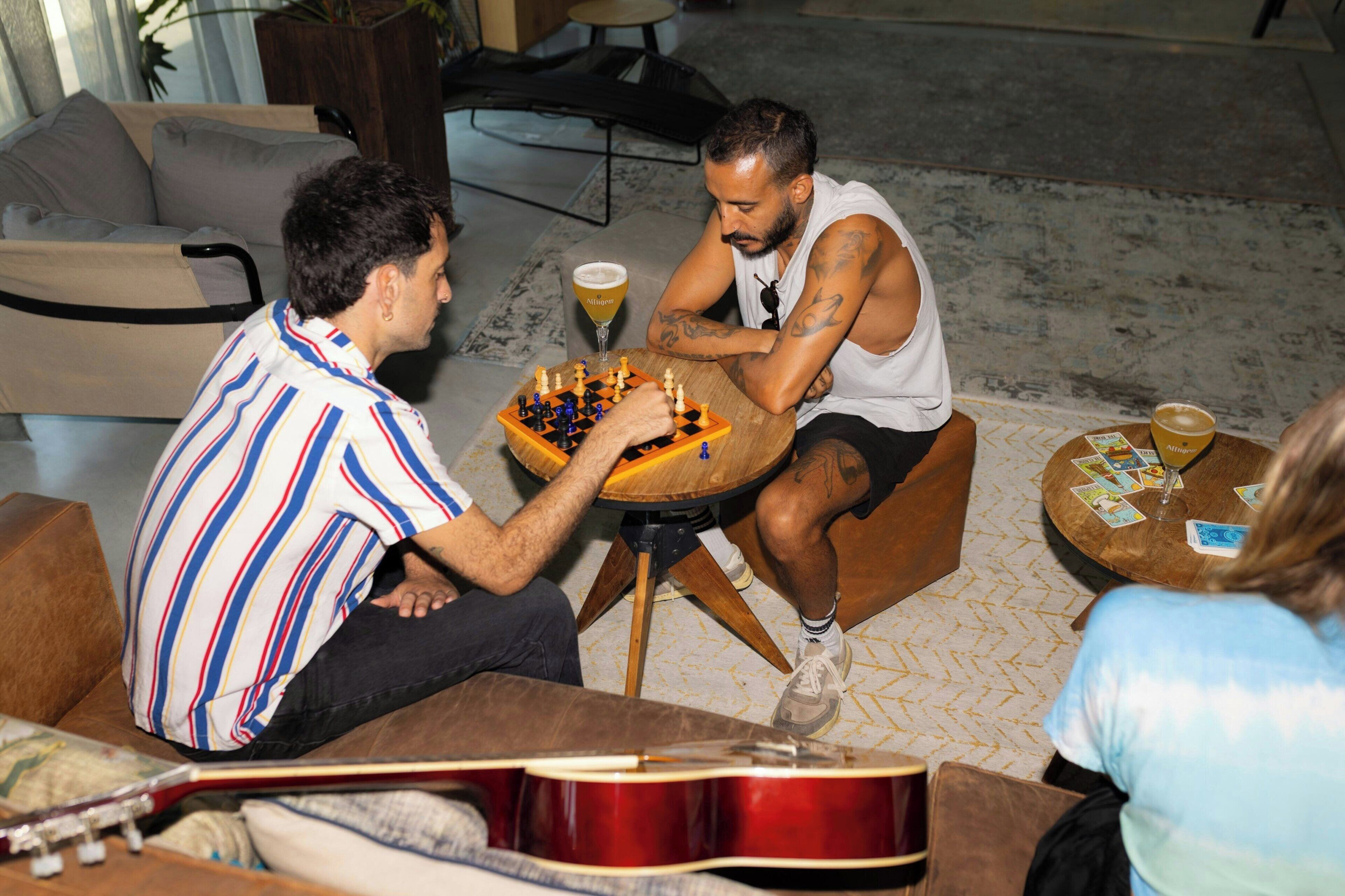 Due persone giocano a scacchi in un ambiente interno informale, con una chitarra e un tavolo rotondo.