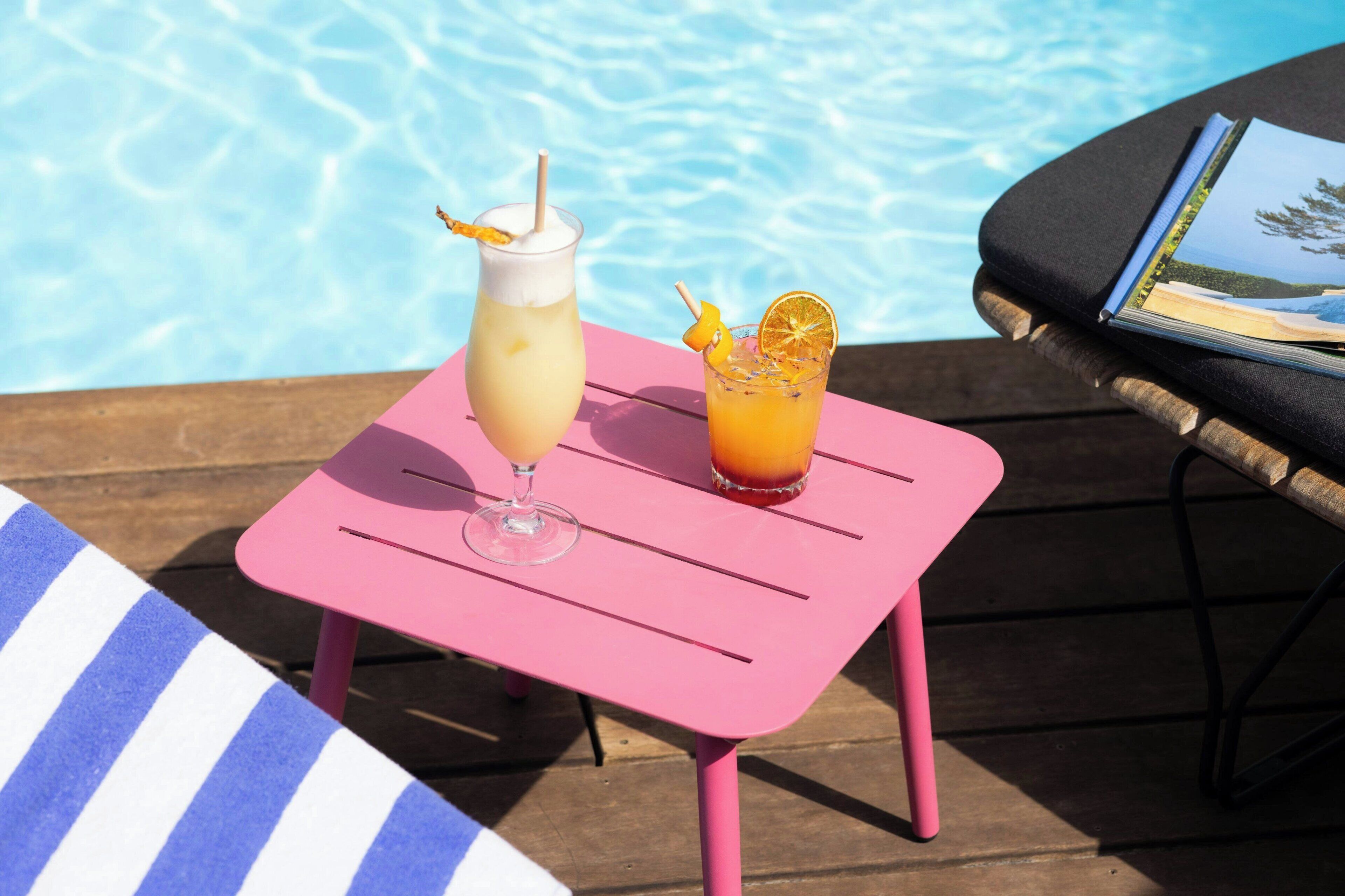 Two drinks on a pink table by a poolside with a lounge chair and a book.