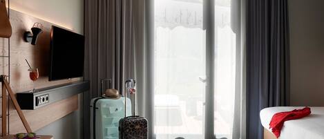 A hotel room with a bed, a desk with a TV, a suitcase, and a window with curtains.