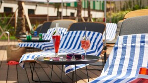 Buitenloungestoelen met gestreepte kussens, een tafel met drankjes en zonnebrillen, en parasols in een zonnige omgeving.