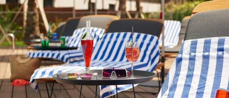 Outdoor lounge chairs with striped cushions, a table with drinks and sunglasses, and umbrellas in a sunny outdoor setting.