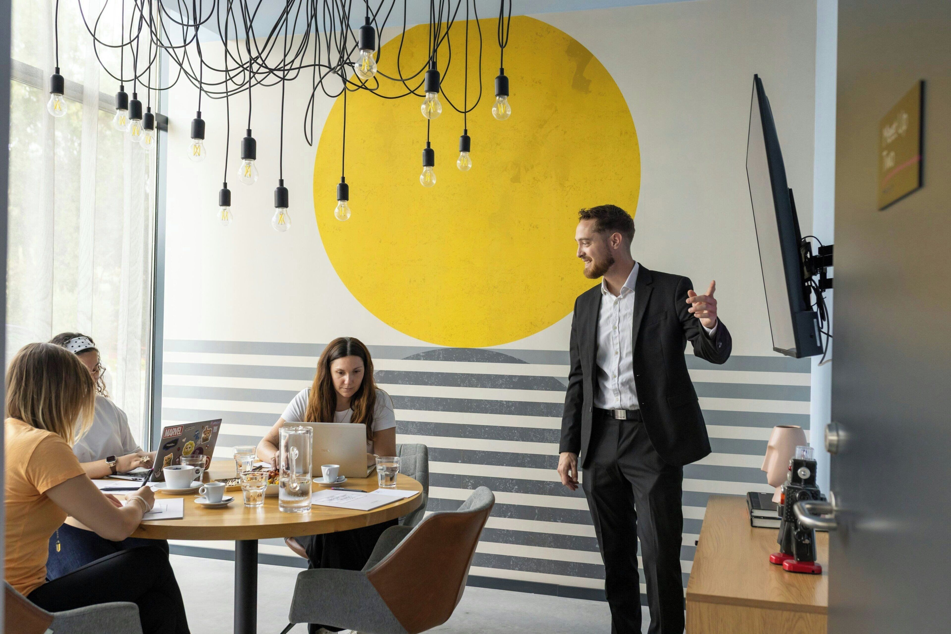 A conference room with a large yellow circle on the wall, a round table with chairs, and a person standing with a laptop.