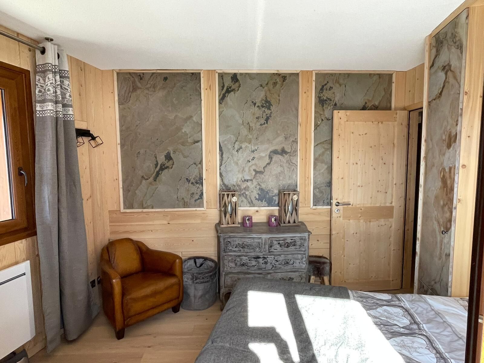 A bedroom with a bed, a wooden dresser, a leather armchair, and a window with curtains.