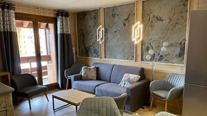 Ein modernes Wohnzimmer mit grauem Sofa, zwei Sesseln, einem Couchtisch und einem Tisch mit Glasplatte. Ein Fenster mit Holzrahmen und Vorhängen, eine Wand mit Marmormuster und ein Kühlschrank sind ebenfalls zu sehen.