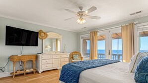 Een slaapkamer met een bed, een televisie, een plafondventilator, een dressoir, een rieten stoel en uitzicht op de oceaan door een deur.