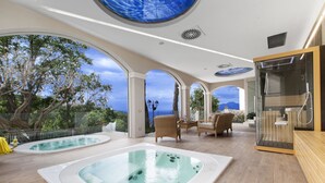 A luxurious indoor pool area with two hot tubs, a glass-enclosed shower, and a seating area with a view of the ocean. - Sorrento Dream Villa (Sorrento)