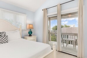 A bedroom with a bed, nightstand, lamp, and a view of a balcony with outdoor furniture. - Tybee Townhome: Ocean Views & Private Path to the Beach (Tybee Island)