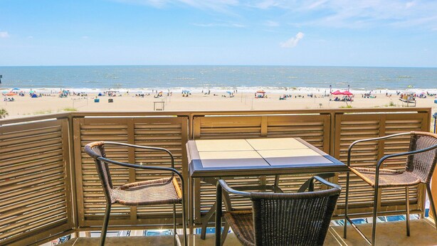 A balcony with wicker chairs and a table overlooking a beach with umbrellas and people. - South Beach Escape | Ocean Views + Walkable Fun (Tybee Island)
