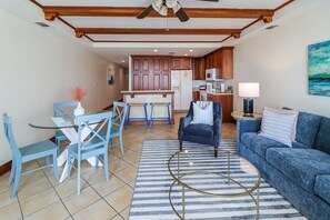 A living room with a blue sofa, a dining table with blue chairs, a kitchen in the background, and a ceiling fan.