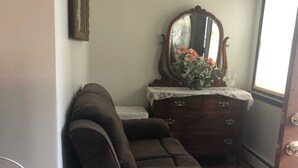 Quarto com sofá marrom, cómoda de madeira com espelho e pequena mesa com um vaso de flores.