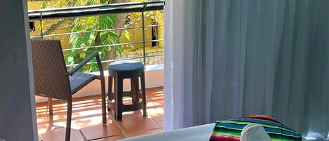 A hotel room with a bed, a chair, a small stool, and a view of a yellow building and greenery through a window.