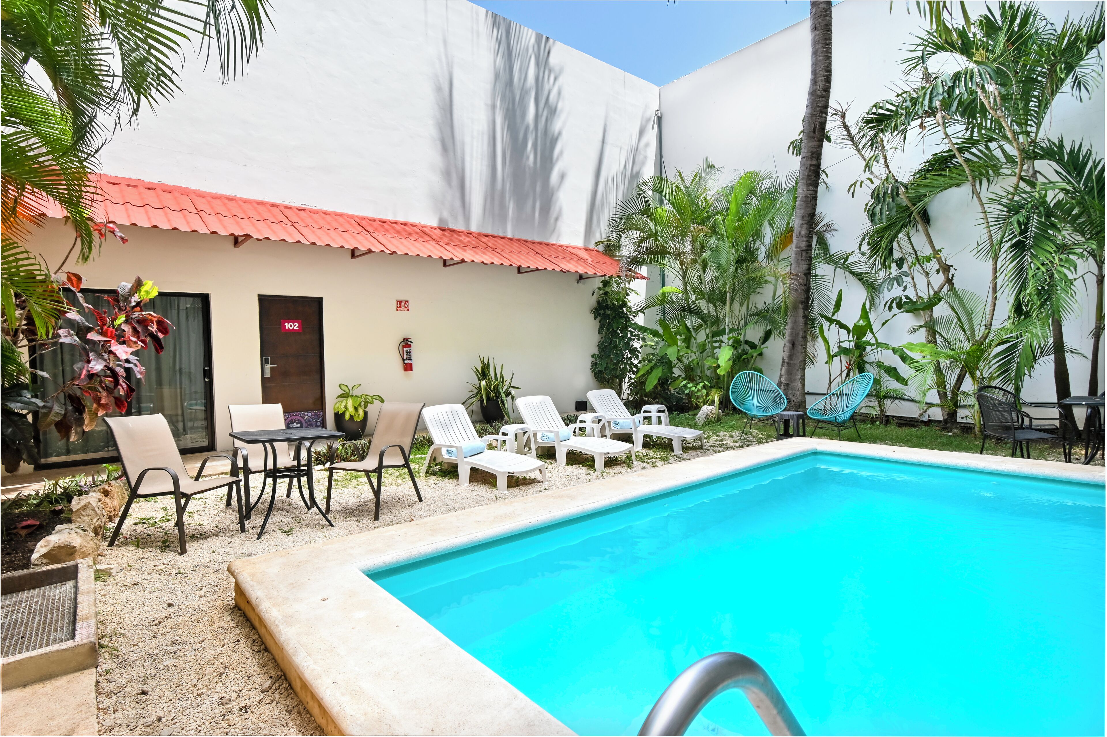 Área de piscina com espreguiçadeiras e uma mesa, cercada por plantas e um edifício com telhado vermelho.