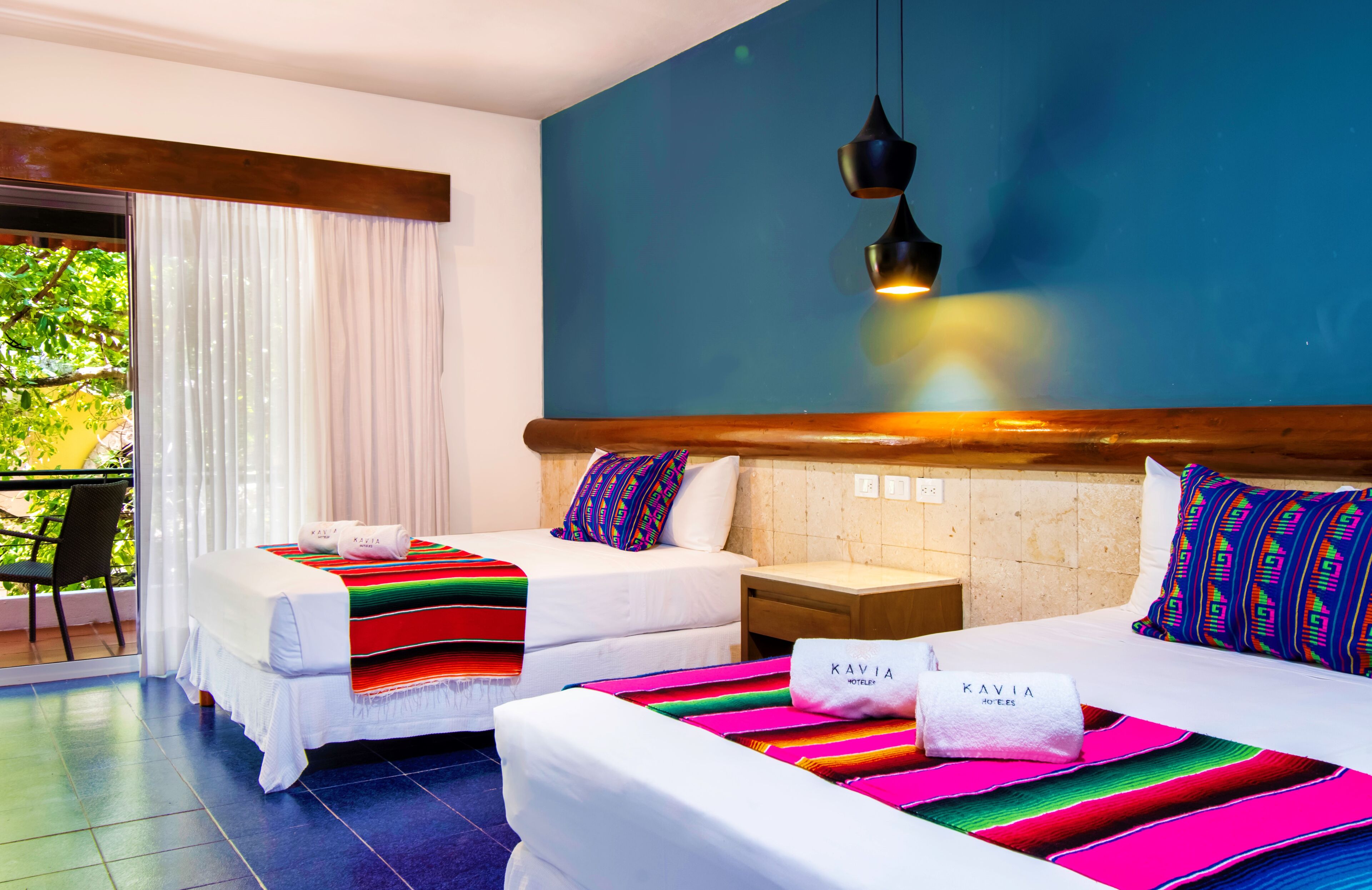 A hotel room with two beds, each with a colorful blanket and a pillow with the hotel name. There is a window with curtains, a wooden headboard, and a hanging light fixture.