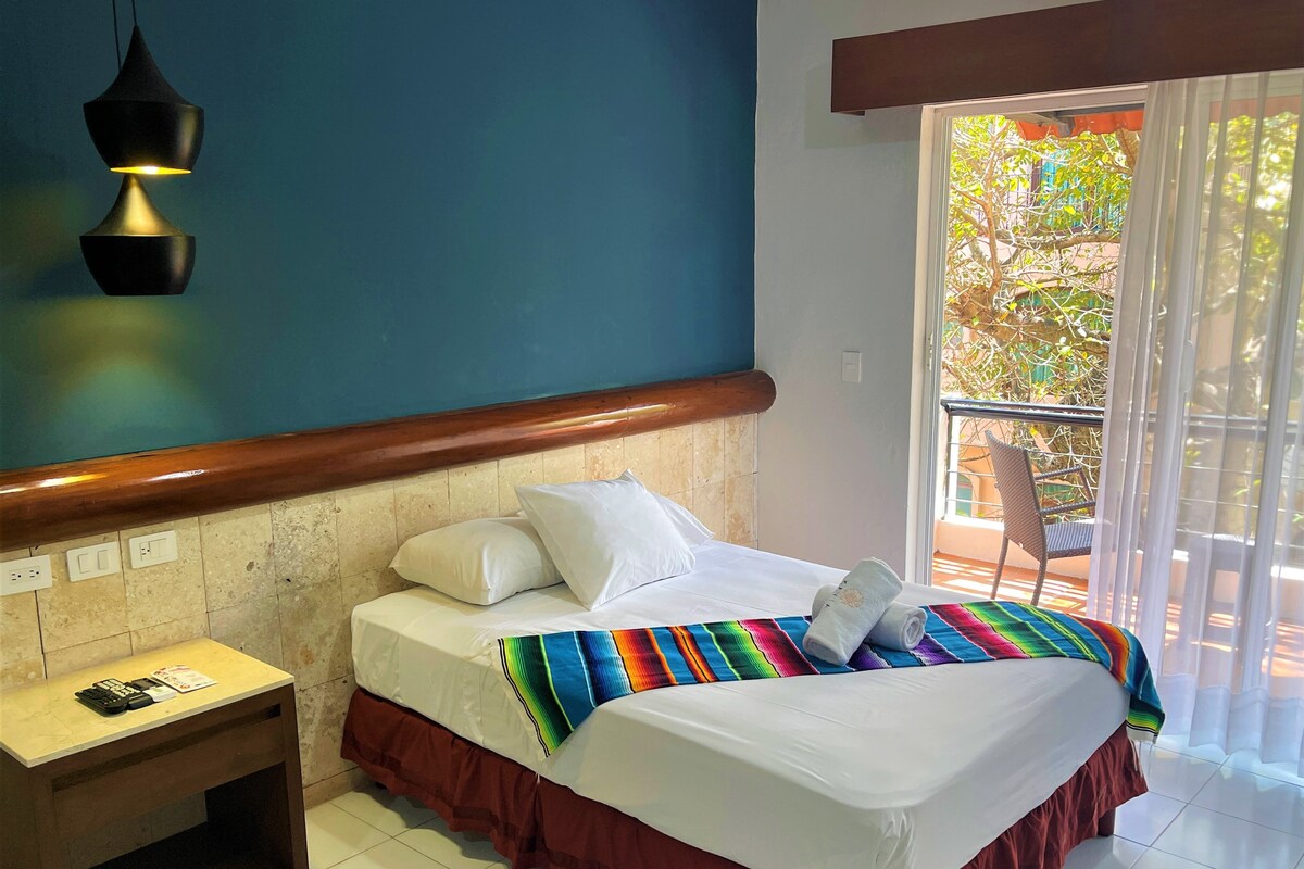 Habitación de hotel con cama, mesita de noche, enchufe y vista al balcón.