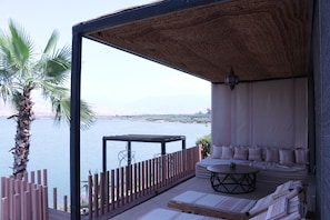 A balcony with a view of a body of water, a palm tree, and a seating area with cushions and a table. - Kalyptus Luxury Camp (Lalla Takarkoust)