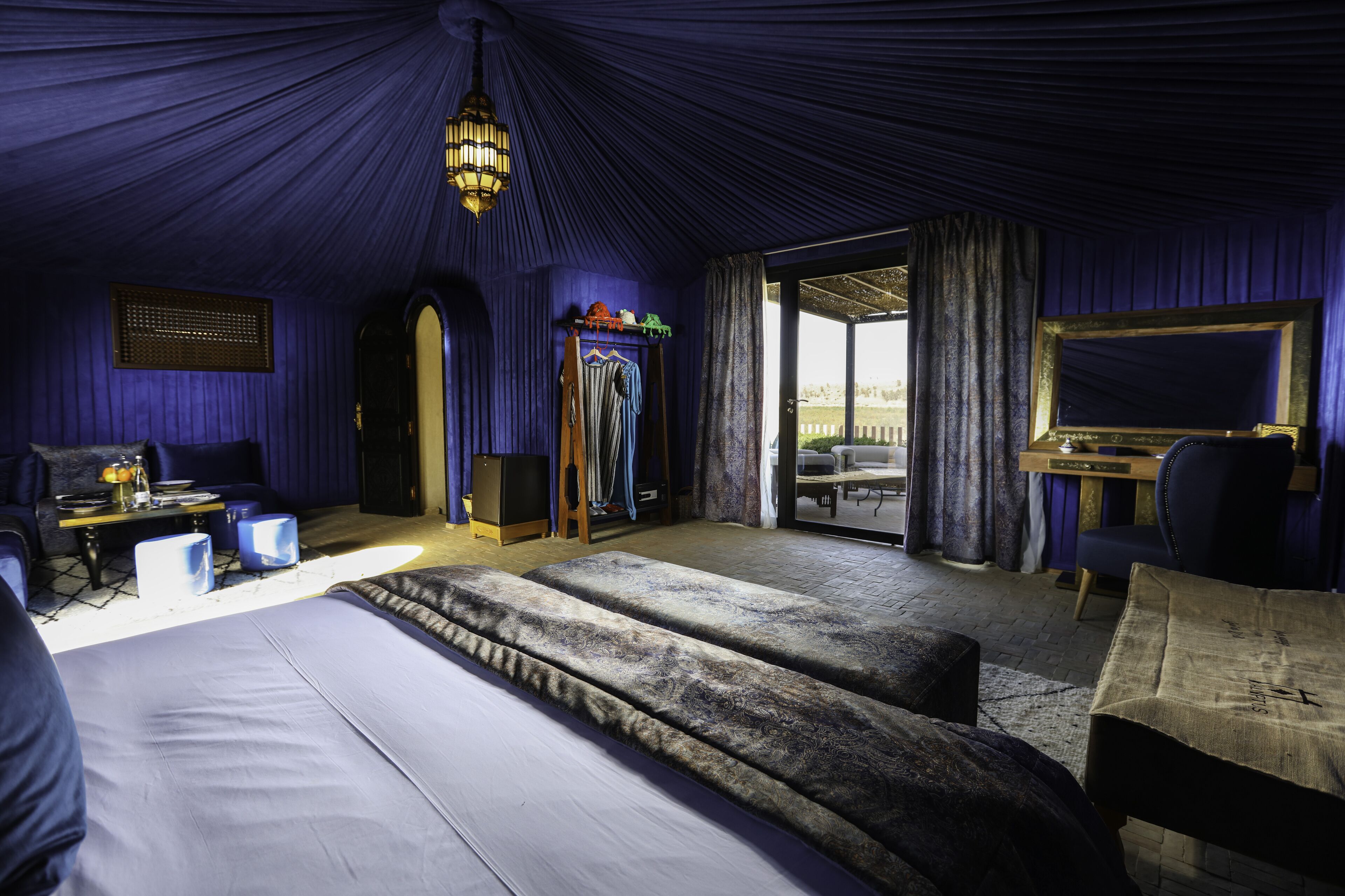 A room with a large bed, a desk, and a chair. There is a window with curtains, a ceiling light, and a door leading outside.