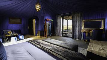 A room with a large bed, a desk, and a chair. There is a window with curtains, a ceiling light, and a door leading outside.