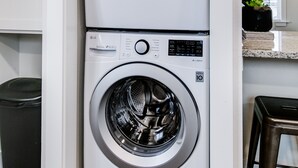 A compact, modern laundry room with two stacked front-loading washing machines, a trash bin, and a bar stool.