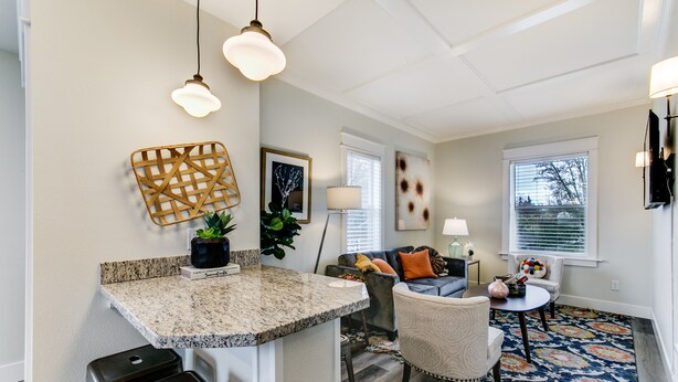 A modern kitchen with granite countertops, a built-in island, and bar stools. There is a living area with a sofa, armchairs, and a coffee table. The room features a patterned rug, wall art, and a large window with blinds.
