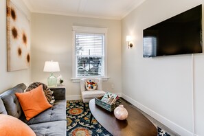 A living room with a grey sofa, a coffee table, a TV mounted on the wall, a window with blinds, and a patterned rug. - The Pine House: Unit A near 6th Ave, UPS, UW, and Point Ruston (Tacoma)