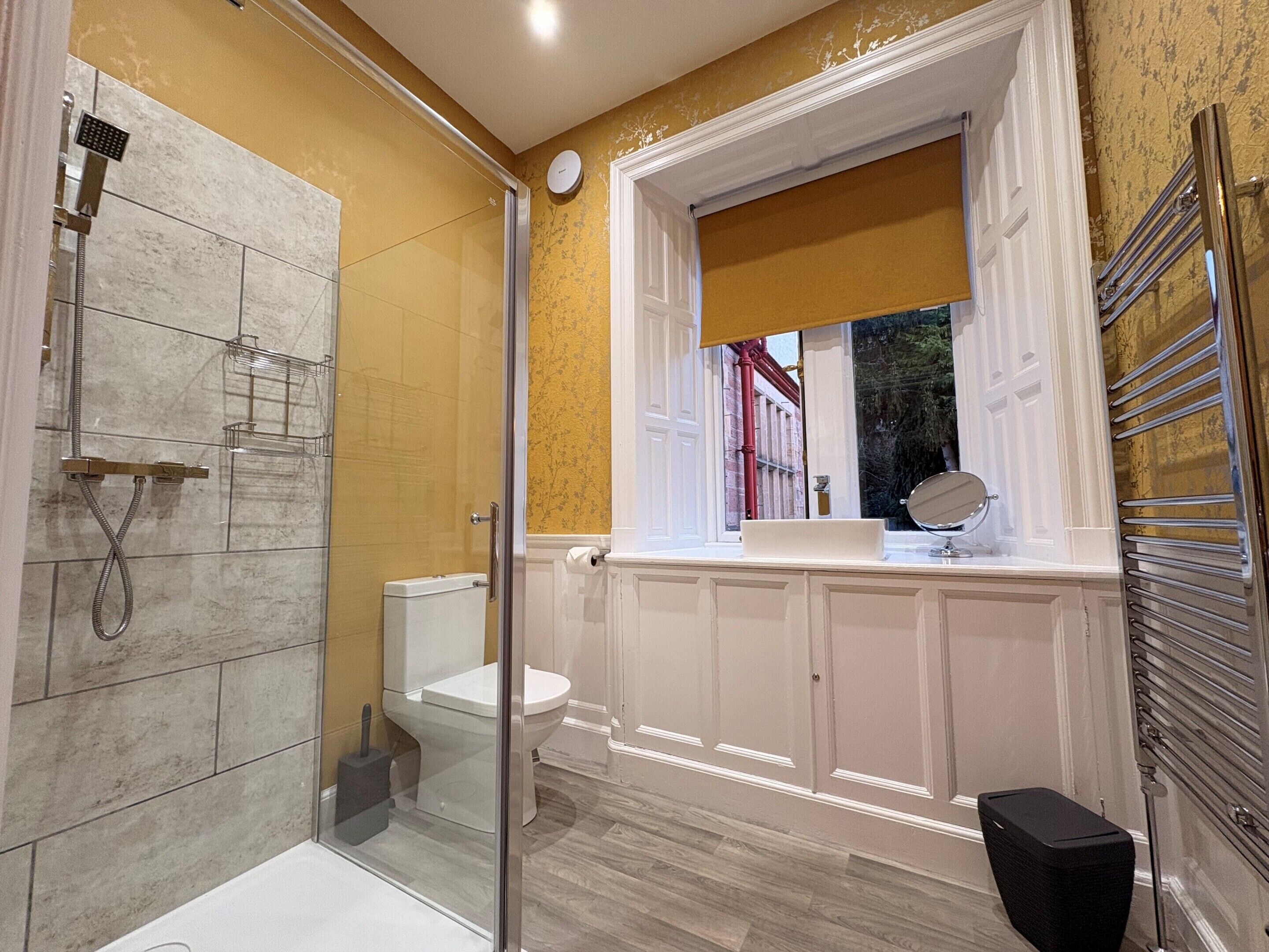 A bathroom with a glass shower enclosure, a toilet, a sink with a mirror, and a window with a view of the outside.