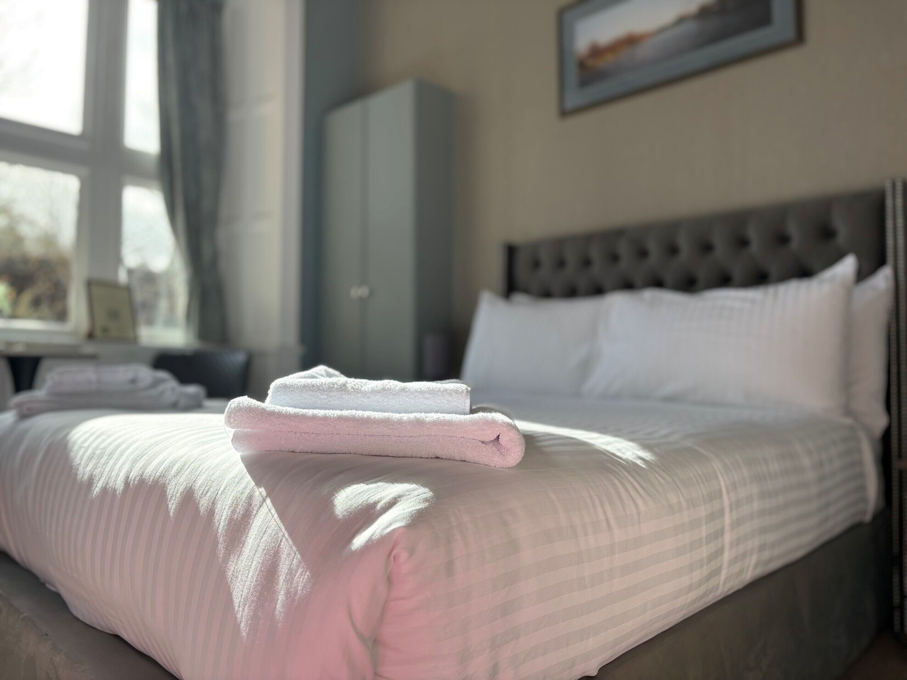 Una cama bien hecha con toallas blancas y rosas, un cabecero gris y una ventana con cortinas.