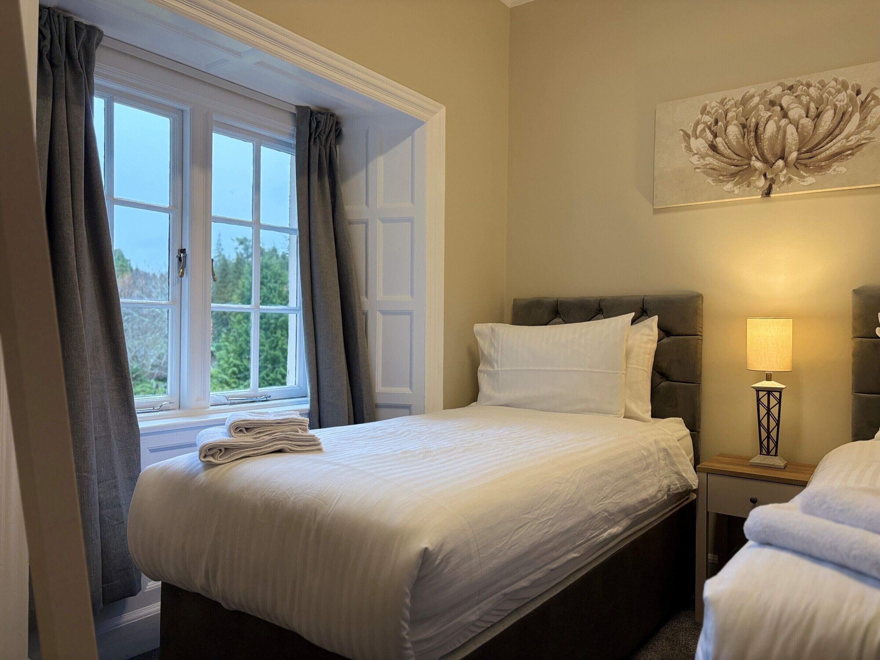 A hotel room with a bed, a nightstand, a lamp, a window with curtains, and a wall-mounted artwork.