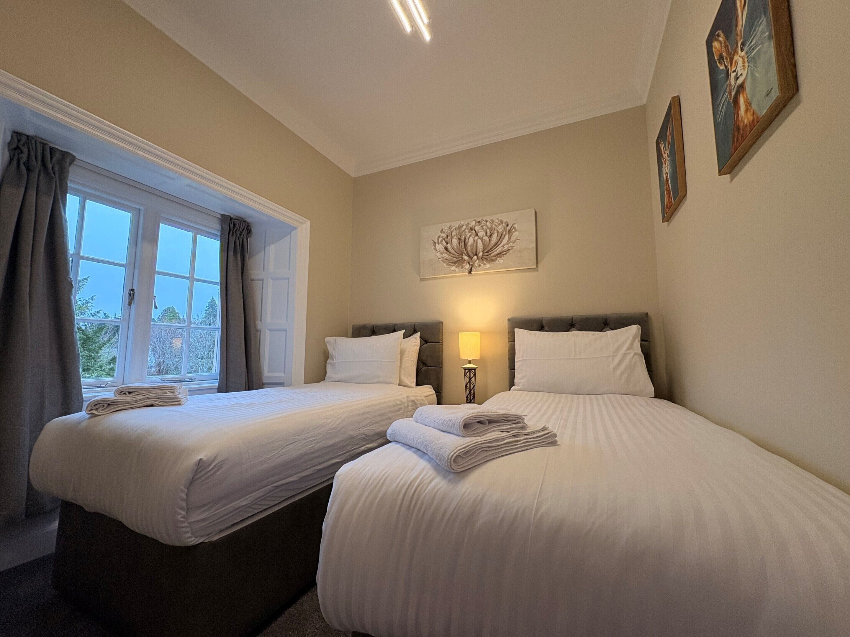 A hotel room with two beds, a window with curtains, a bedside lamp, and a painting on the wall.