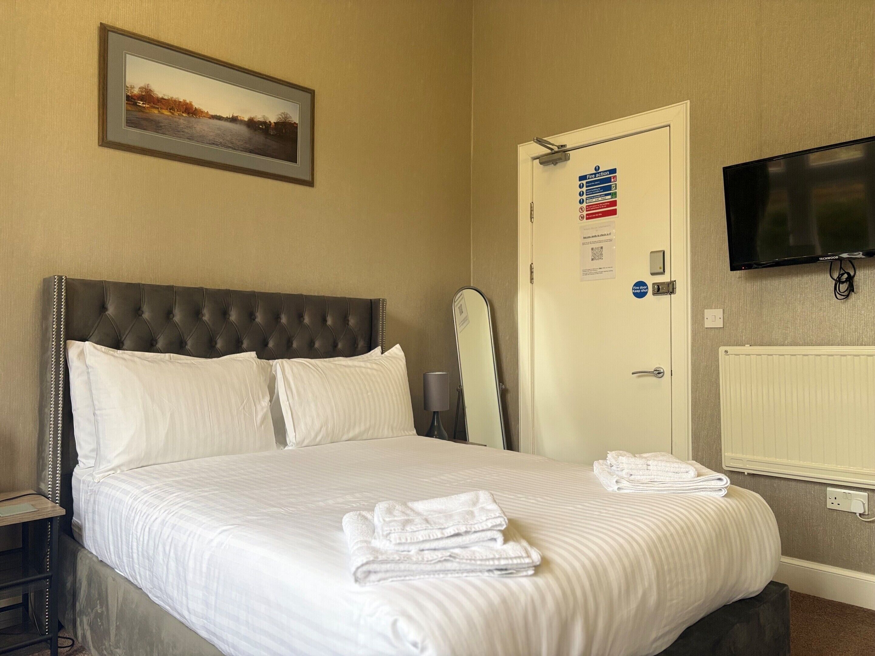 Une chambre d’hôtel avec un grand lit, un tableau encadré au mur, une porte avec un panneau indiquant les consignes de sécurité en cas d’incendie et un téléviseur fixé au mur.