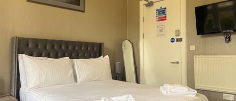 Une chambre d’hôtel avec un grand lit, un tableau encadré au mur, une porte avec un panneau indiquant les consignes de sécurité en cas d’incendie et un téléviseur fixé au mur.