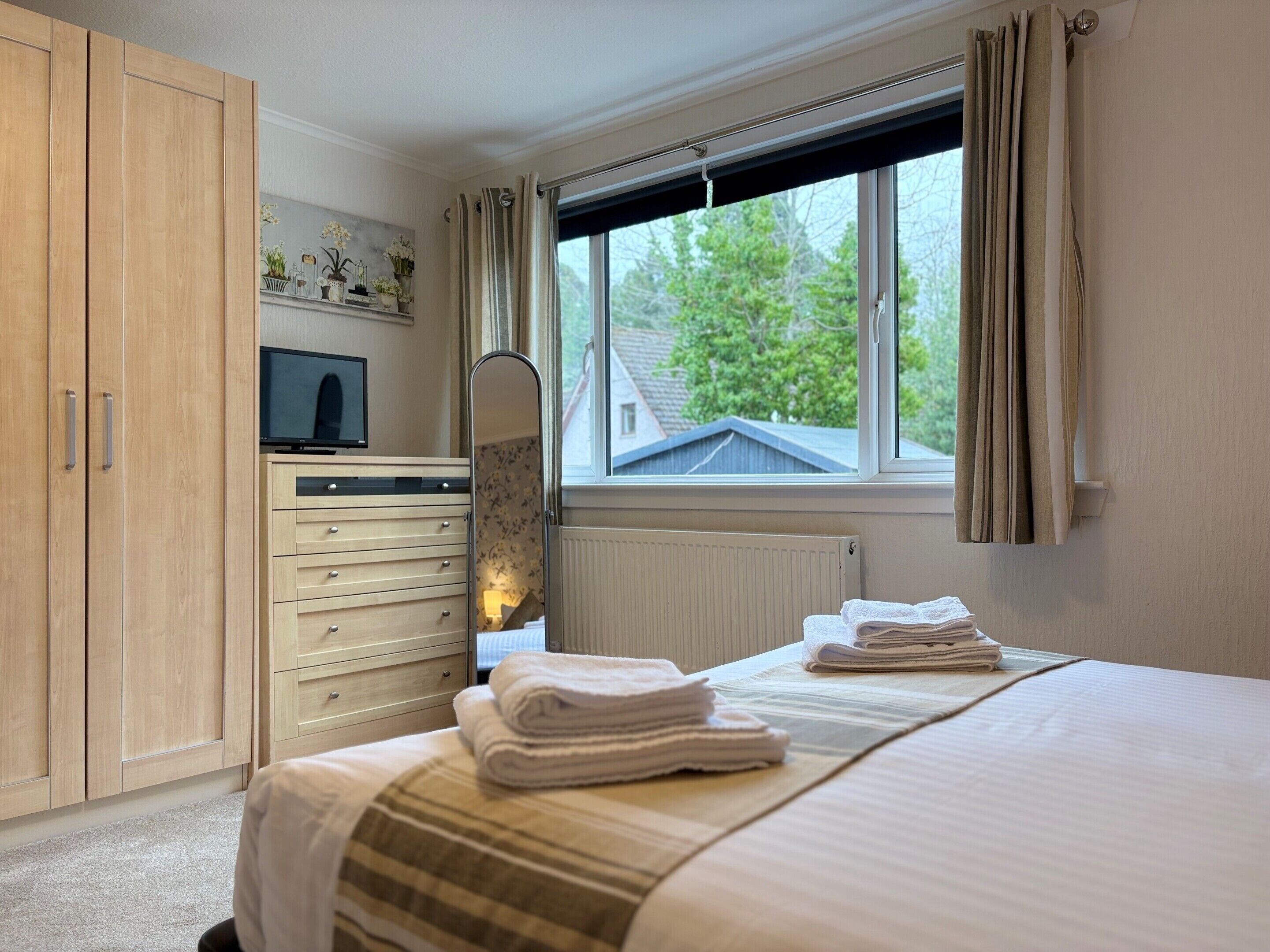 Een slaapkamer met een bed, een dressoir, een televisie, een raam met gordijnen en een radiator.