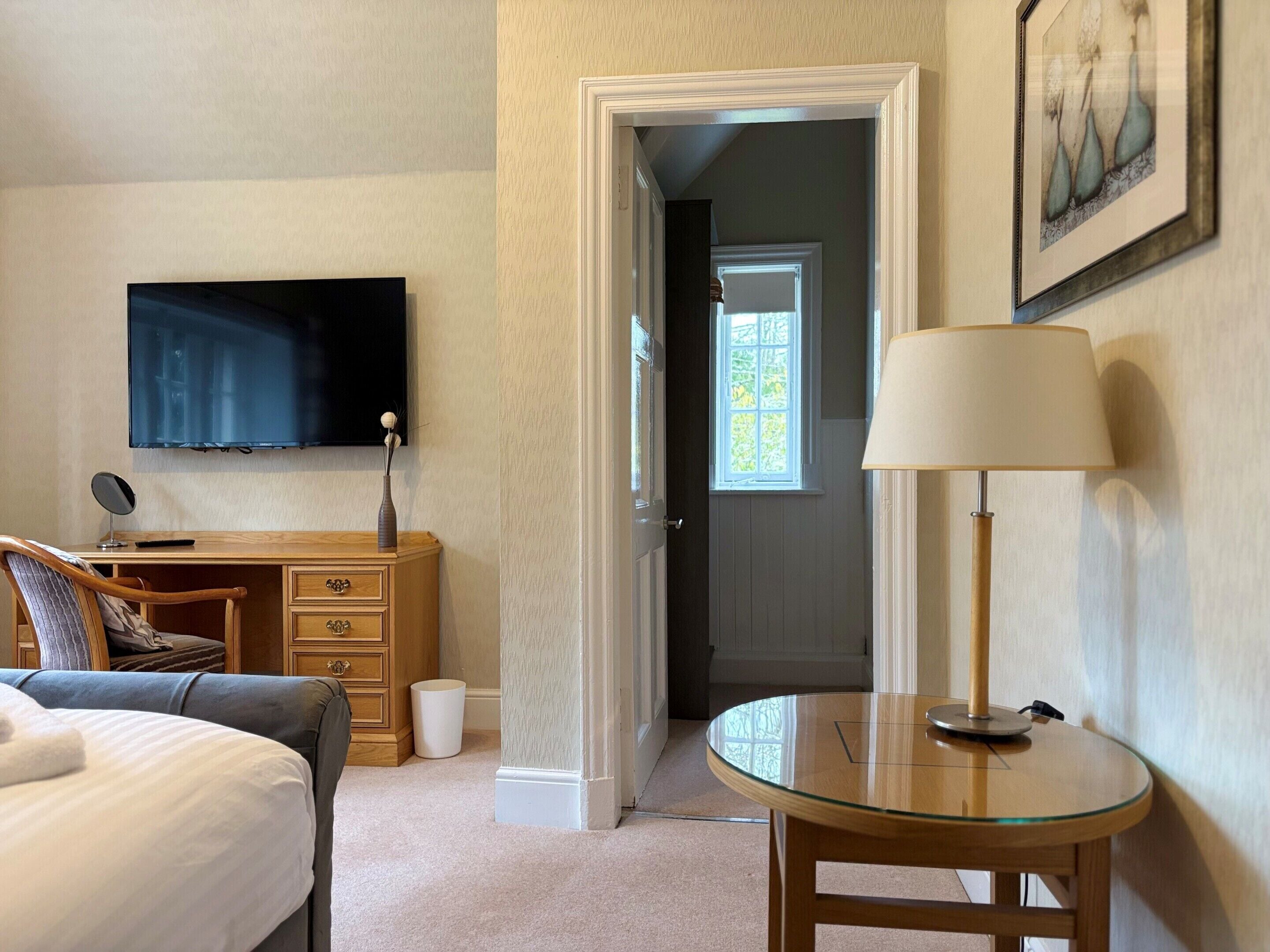 Una camera d'hotel con un letto, una scrivania con un computer, una televisione, una lampada e una porta che dà su un'altra stanza.