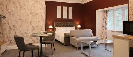 A hotel room with a bed, a grey armchair, a round table with a lamp, a television, and a window with curtains.