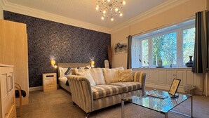 Un dormitorio con cama, sofá, una mesita de cristal, cómoda y ventana con vista a árboles.