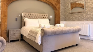 Un dormitorio con un cabecero capitonado, una gran estructura de madera arqueada, una cama con ropa de cama blanca, una mesita de noche y un espejo en la pared.