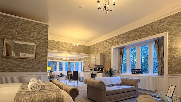 Habitación de hotel con una cama grande, un sofá, un comedor y vistas a árboles por la ventana.