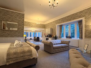Ein Hotelzimmer mit einem großen Bett, einer Couch, einem Essbereich und Blick auf Bäume durch das Fenster.