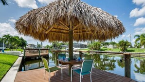 A wooden deck with a thatched umbrella, two chairs, and a table by a calm water body, surrounded by greenery and a clear sky.