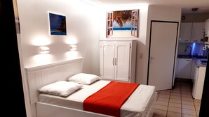 Un dormitorio con cama, paredes blancas, una colcha roja y un cuadro de una playa sobre la puerta.
