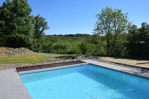 A rectangular swimming pool with a wooden deck edge, surrounded by a concrete border, set in a landscaped area with trees and a grassy hill in the background.