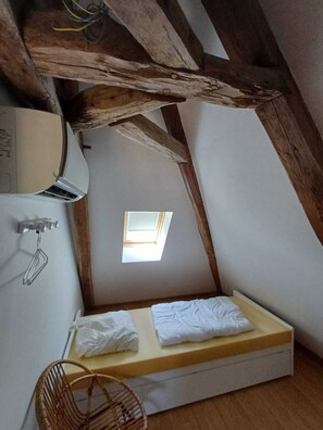 A small, attic-style bedroom with a sloped ceiling, a skylight, a bed with white linens, a wicker chair, and a wall-mounted air conditioning unit.