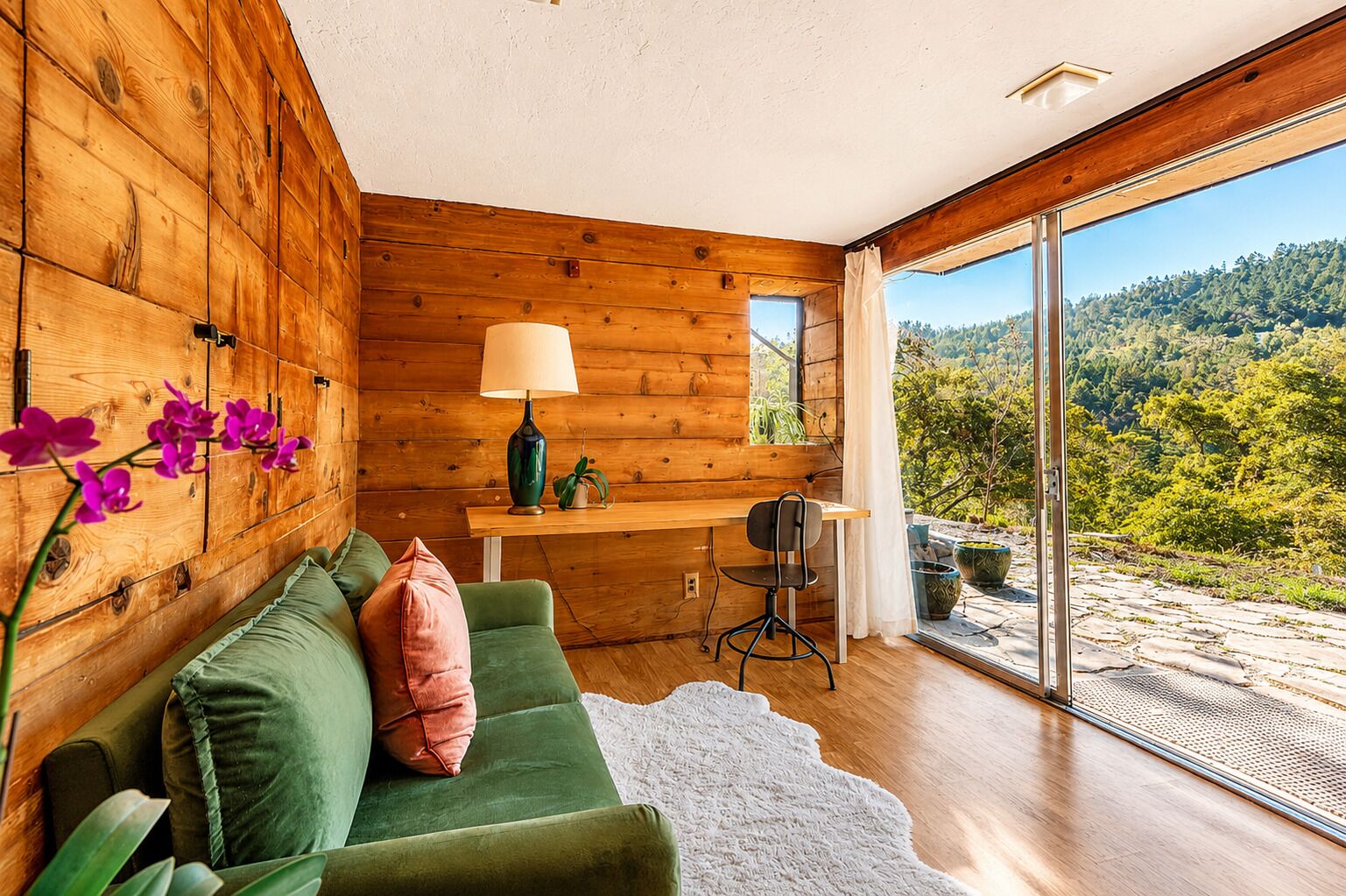 A modern interior with a wooden wall, a green sofa, a lamp, and a view of a forested landscape through large windows.