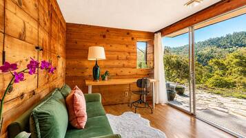A modern interior with a wooden wall, a green sofa, a lamp, and a view of a forested landscape through large windows.