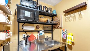 Une cuisine compacte équipée d’un four à micro-ondes, d’une bouilloire et d’un évier en acier inoxydable. Des étagères au-dessus de l’évier servent à ranger divers ustensiles de cuisine, et un porte-verres suspendu permet d’y accrocher des verres à vin.