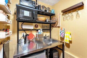 A compact kitchen with a microwave, kettle, and stainless steel sink. There are shelves above the sink holding various kitchen items and a hanging rack with wine glasses. - Enchanted Waterfall Sanctuary (Mill Valley)